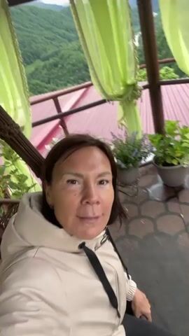 A woman is sitting in a macrame swing inside a gazebo, filming herself. She is wearing a light beige hooded jacket over black clothing and has shoulder-length brown hair. The gazebo has a red roof, green curtains, and potted plants around the edges. The background shows a view of mountains and trees. The camera is positioned at eye level, and the woman looks directly into it. The swing moves slightly.