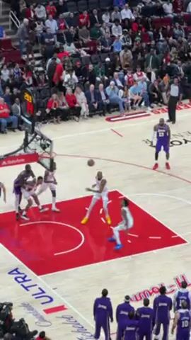 The video shows a basketball game. Two teams are playing. One team wears white and red jerseys and the other team wears purple and yellow jerseys. The players are running around the court. They are passing the ball and dribbling. A referee is standing on the court, watching the game. There are fans in the stands. The court has red and blue symbols on it. The video shows a basketball game.