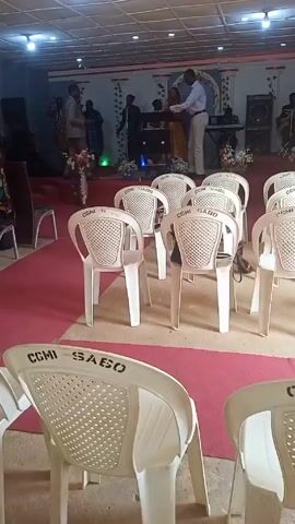 The video shows an interior view of a building, likely a church, with several white plastic chairs in the foreground. The chairs have the inscription "CGMI SABO" on them. A red carpet covers the central aisle leading to a raised stage area at the far end. On the stage, a band is performing, and a couple is standing close together, seemingly being prayed over or blessed by one of the band members. Several other individuals are present on stage, including musicians and singers. The lighting in the room is relatively dim, with overhead fluorescent lights and additional stage lighting. The overall atmosphere appears to be that of a religious gathering or celebration.