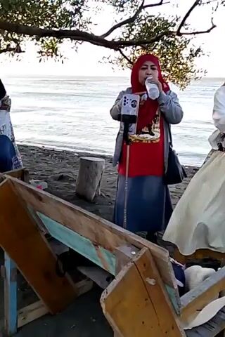 Two women stand on a beach. The woman on the left wears a red hijab and a red t-shirt over a blue skirt. She is drinking from a water bottle and holding a phone on a selfie stick. The woman on the right wears a light-colored hijab and a white t-shirt with a graphic print, and a long, light-colored skirt with a patterned panel. She is holding her hands near her face and appears to be speaking or eating. Behind them is a body of water, likely the ocean, with waves. The ground is sand and there are some logs and a wooden structure in the foreground. A tree with branches overhangs the scene. The lighting suggests it is either early morning or late afternoon.