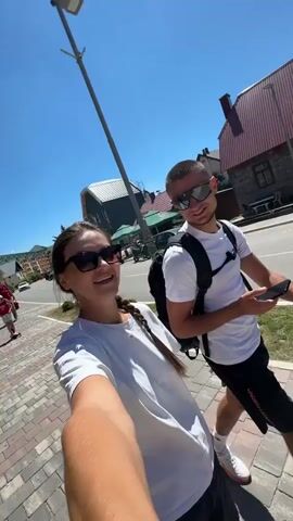 Two young adults, a woman and a man, are taking a selfie outdoors on a sunny day. The woman is in the foreground, holding the camera and looking at the man. She has dark hair pulled back and is wearing a white t-shirt and sunglasses. The man is standing next to her, also wearing sunglasses and a white t-shirt with a black backpack. He is looking at the camera and holding a smartphone. They are on a paved walkway next to a road. In the background, there are buildings with red roofs, trees, and a blue sky.