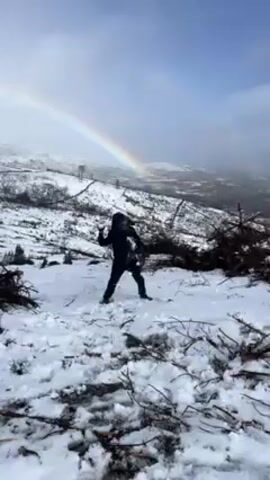 Two people in black coats engage in a snowball fight in a snow-covered mountainous landscape. A vibrant rainbow arcs across the sky in the background, with the lower part of the scene cluttered with branches and debris. The sky is partially overcast, with a visible vista beyond the snow-covered landscape.