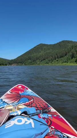 A first-person perspective shot from a paddleboard on a body of water. The front of the paddleboard is visible, decorated with a red, blue, and white koi fish design and the word "KOE." A blue bag and a pair of white sandals are on the paddleboard. The water is dark with ripples. In the background, a large, tree-covered hill rises under a clear blue sky. The scene appears to be a scenic outdoor environment.