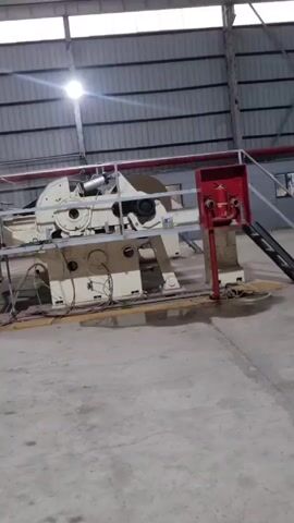 The video shows a piece of heavy machinery in a large industrial space. The machinery is tan and white with exposed wires and metal supports, including a large circular blade. A red fire safety station is located near the machine. The space has a concrete floor and a corrugated metal ceiling with a single bright light overhead.