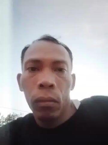 A close-up, low-angle video captures a man's face from the chest up. He has short, dark hair, a receding hairline, and prominent cheekbones. He wears a black t-shirt. The background is out of focus, showing a bright sky and some indistinct greenery. The man's gaze is directed forward, and his expression is neutral. The video is short, with no discernible action or interaction.