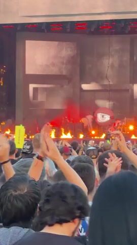 The video captures a concert scene from behind a crowd. The stage is set with a large screen displaying abstract, monochromatic shapes. Flames erupt from the stage floor in front of the performers and their instruments. Several band members are visible on stage, including a drummer and a figure with a microphone. The crowd, composed of many individuals, has their hands raised, suggesting engagement with the performance. The lighting is dramatic, with red hues from the flames and stage lights casting a warm glow. The overall atmosphere appears energetic and exciting.