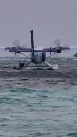 The provided video shows a seaplane on the water, with a person next to it and a red buoy nearby. The seaplane is white and blue, has two propellers, and a red light on its tail. The water is choppy and the sky is overcast. The person appears to be working near the seaplane. The background shows a distant landmass.