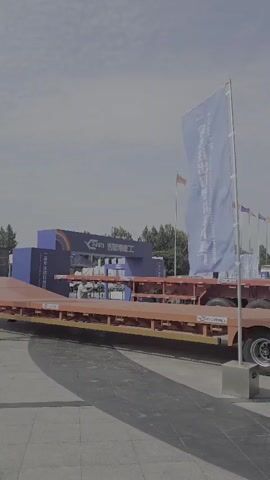 The video pans from left to right across a display of large trailers and trucks parked outside a modern convention center with a glass facade. A tall blue flag with Chinese characters stands in the foreground to the left of the frame. The trailer on the left is an orange flatbed trailer with multiple axles and large black tires. To its right is a silver tanker trailer. Further to the right, another silver trailer, this one a dump truck, is parked. Two people are visible walking near the dump truck in the middle ground. In the foreground to the right, a woman in a red shirt and glasses walks towards the camera, holding a cell phone. The convention center building has a prominent sign with Chinese characters and smaller English text that reads "LIANGSHAN INTERNATIONAL EXHIBITION CENTER" and "THE 22ND CHINA (LIANGSHAN) SPECIALPURPOSE VEHICLE EXHIBITION". Several smaller flags are visible further back in the scene. The sky is overcast.