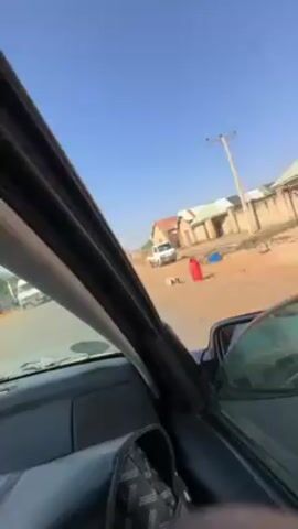 The video shows a view from inside a car moving down a red dirt road. The sky is clear and blue. Buildings can be seen to the right of the road, and people and vehicles appear to be on a paved road to the left. Power lines run along the right side of the dirt road.