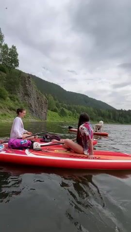 The video shows a group of people paddleboarding on a river. In the foreground, two women are seated on separate red paddleboards. The woman on the left, wearing a white hoodie and purple shorts, is holding a paddle and looking towards the camera. The woman on the right, wearing a colorful patterned shirt, is facing away from the camera. Behind them, another paddleboard is visible with a person and a small white dog on it. The paddleboards are on calm, dark water. The background features a steep, rocky cliff face on the left, covered in green vegetation. Further in the background, a lush, tree-covered mountain range stretches across the horizon under a cloudy sky. The water reflects the sky and surrounding landscape.