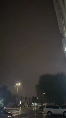 The video depicts a dark, stormy night with flashes of lightning illuminating the sky. In the foreground, a street lamp shines brightly, casting light on the lower portion of the frame. Adjacent to the street lamp is a silhouette of a tree, its leaves indistinguishable in the darkness. In the distance, there's a row of buildings, partially visible under the dim lighting. Cars are parked along the street, and a red light can be seen in the background. The main focus of the video is the sky, which is filled with dark, ominous clouds interspersed with frequent flashes of lightning.