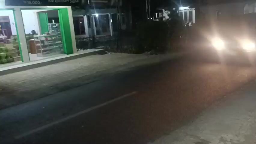 A black vehicle with its headlights on is driving down a paved road at night. To the left of the road, there is a storefront with a green trim and a display of goods inside. The storefront appears to be a shop of some kind. The road has a white line marking the lane. The overall environment is dark and appears to be a street in a town or village. The video is short and the vehicle is the main subject.