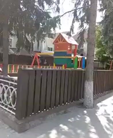 The video shows a playground. In the foreground, there is a wooden fence. Behind the fence, there is a colorful playground structure with a red roof, blue and green platforms, and yellow and red supports. To the left of the structure, there is a red swing set. In the background, there are trees and buildings. There are also benches visible to the right of the playground. The ground of the playground is covered with sand or wood chips. The overall scene suggests a sunny day at an outdoor playground.