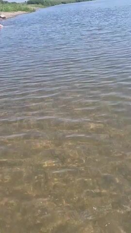 The video shows a view of a body of water from a low angle, appearing to be shallow near the shore. The water is clear enough to see the bottom, which is composed of sand and small rocks. The surface of the water has small ripples. In the distance, on the left side of the frame, a person is visible in the water. The shoreline is visible in the background, with some sparse vegetation. The sky is blue and there are some clouds.