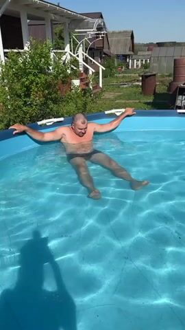 A man wearing swimming trunks is lying in a blue above-ground pool outdoors. He stretches his arms out to the sides. There are houses and trees in the background. The man looks at the camera, then looks off to the right, and then looks back at the camera.