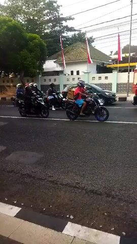 The video shows a group of motorcycles riding on a paved road. There are several people on the motorcycles, most of whom are wearing helmets. One motorcycle in the foreground has a rider with blue shorts and a red helmet. Another motorcycle has a wooden box attached to the back. In the background, there is a building with white walls and a tiled roof. Two Indonesian flags are visible, one on a pole and one attached to the building. There is also a black car and some trees. The road appears to be in a residential or commercial area. The lighting suggests it is daytime, possibly late morning or afternoon.