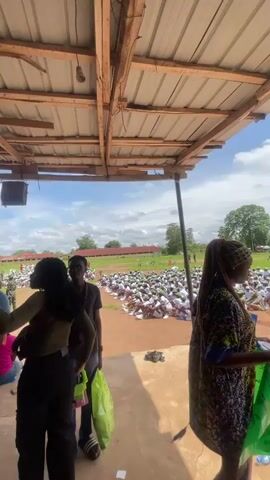 The video shows a group of people under a covered structure, looking out at a large gathering of people sitting on the ground in an open field. The crowd in the field appears to be mostly young people, possibly students, dressed in white. There are trees and buildings visible in the background, under a cloudy sky. In the foreground, under the structure, are several individuals. Two women and a man are standing in the center, engaged in conversation. One woman is holding a pink folder, and the other is holding a green bag. Another person, a woman with braided hair, is partially visible on the right. The atmosphere seems casual, with people observing the event in the field. The structure is made of corrugated metal and wood. The ground is dirt and concrete.