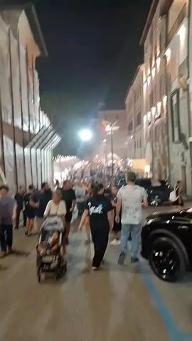 A nighttime street scene shows a crowd of people walking down a narrow, cobblestone street lined with buildings. The buildings have lights on, and some have scaffolding. The street is partially blocked by parked cars, and there are temporary structures, possibly for an event or market, further down the street. The crowd is diverse, with people of various ages and ethnicities. Some individuals are walking with strollers, while others are walking in groups or alone. There are streetlights and bright lights from the event area illuminating the scene. The overall atmosphere appears to be festive and lively.