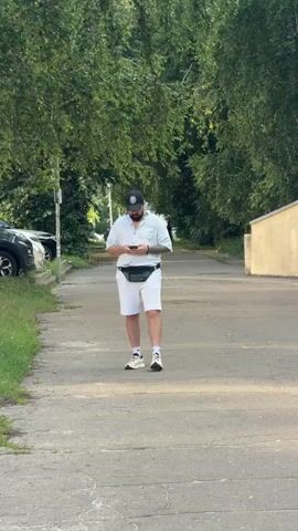 A man wearing a white short-sleeved button-up shirt, white shorts, a black baseball cap, and white sneakers walks towards the camera on a paved sidewalk. He is holding a smartphone in his hands and looking down at it. A dark green fanny pack is worn across his waist. He has a beard and tattoos are visible on his left arm.

The sidewalk is bordered by green grass and trees on the left and a textured beige wall on the right. Several cars are parked on the left side of the path. In the background, more trees and foliage create a lush, green canopy overhead. A few other people are visible walking in the distance on the sidewalk. The lighting suggests it is daytime, likely late afternoon or early evening, with the sun casting shadows. The overall scene depicts a casual stroll in a park-like or suburban setting. There is no visible text in the media.