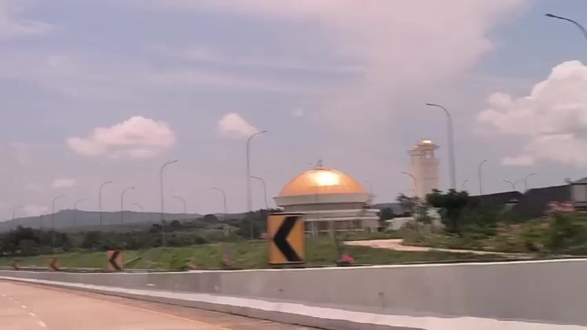 The camera travels along a highway in what appears to be a tropical region. The sky is partially cloudy, and there are green hills in the distance. The road is bordered by a concrete barrier and grass with directional arrows painted on the barrier. There are lampposts along the side of the road. Various buildings are visible in the background, including a mosque with a gold dome and what appears to be a parking area or rest stop with huts. Signage directs drivers to either a "Parkir Kendaraan Besar" (Big Vehicle Parking) or "Parkir Kendaraan Kecil" (Small Vehicle Parking) area.