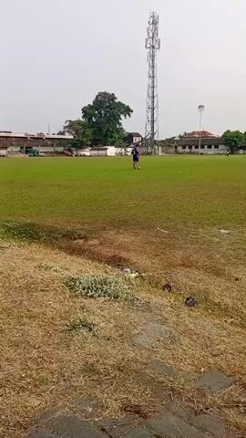 A person is playing soccer on a grassy field. There is a large tower in the background, along with some buildings and trees. The sky is overcast.