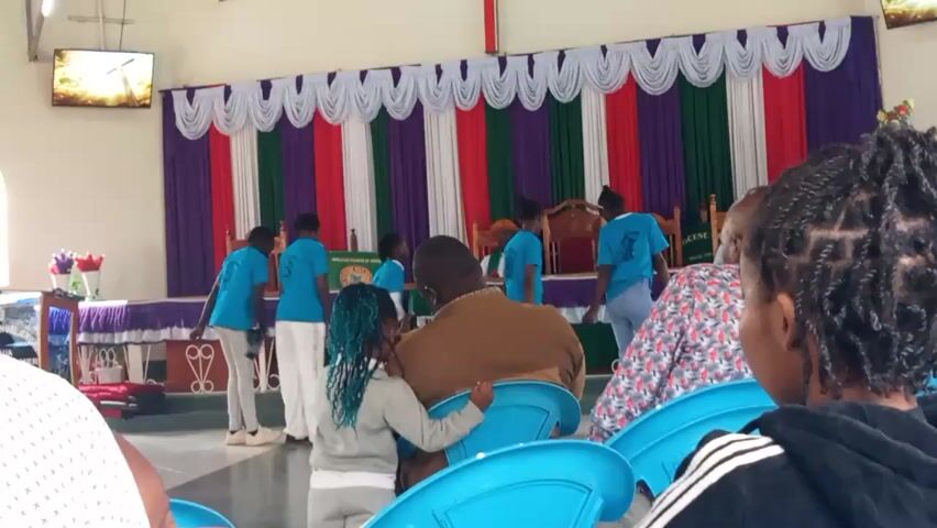 A group of children in matching blue t-shirts are standing in a church in front of a colorful curtain backdrop. Some of the children are looking at the camera while others are looking away. A woman with blue hair is standing in the foreground looking at the children. An adult man is sitting down in the foreground, facing away from the camera. There are also other people sitting in the audience in the foreground, also facing away from the camera. The room appears to be a church or similar place of worship, with chairs and a stage. There is a television screen mounted on the wall in the background.