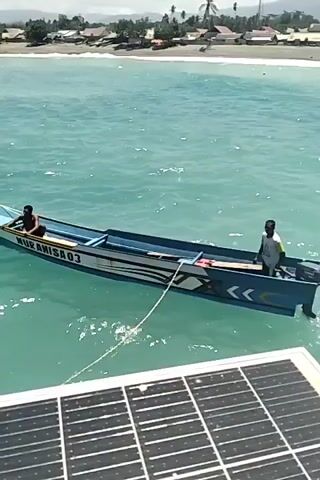 A blue and white motorboat named MURAMISA03 with two men on board is tethered to a solar panel. One man is seated in the front of the boat, and the other is standing at the back, operating the motor, which is creating a wake in the turquoise water. In the background, a coastline with buildings and vegetation is visible under a clear sky.