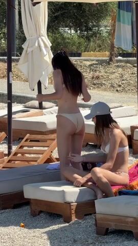 Two young women are on a beach. One woman, wearing a light-colored bikini, is bent over and adjusting herself. The other woman, wearing a white bikini and a gray baseball cap, is sitting on a lounge chair, looking down at her hands. The background shows more lounge chairs, beach umbrellas, and a sandy, rocky area with some dry vegetation. The scene is brightly lit, indicating it's daytime.