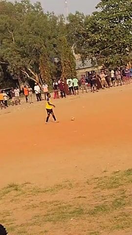 A medium shot shows a group of people playing soccer on a dirt field surrounded by a crowd of spectators. The field is a reddish-brown color and there is grass around the perimeter. The trees are green and lush. The crowd consists of people of various ages and ethnicities, dressed in casual clothing. A player in a yellow and black jersey is dribbling the ball towards the goal. Other players in white and black jerseys are running alongside him. The camera is handheld and slightly shaky. The overall impression is of a local soccer match being played in a community setting.