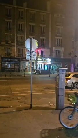 A man rides a green city bike down a quiet city street at night. The street is lined with buildings with evenly spaced windows and a few storefronts. There is a parking meter on the sidewalk next to a parked white car. A sign with street directions is visible above the parking meter. After the bicyclist passes, another man walks along the sidewalk, putting something in his mouth.