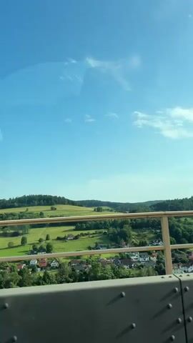 The video is a horizontal, panning shot of a rural landscape, taken from a vehicle. In the foreground, a grey metal railing with rivets runs along the bottom of the frame, indicating the perspective is from a bridge or elevated roadway. A lighter-colored metal railing with vertical posts and horizontal bars is visible in the mid-ground, separating the viewer from the scene beyond. The background features rolling green hills covered in lush trees and fields. A small village with houses and scattered trees is nestled within the valley. The sky is a clear, bright blue with a few wispy white clouds. The sun appears to be high in the sky, casting a bright light on the landscape. The overall impression is of a sunny, serene day in the countryside.