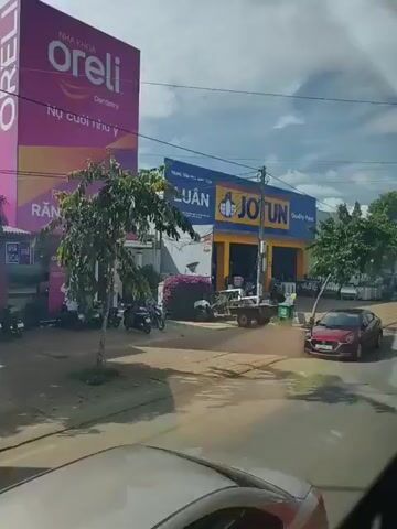 The video shows a street view with several buildings and vehicles. In the foreground, a red car is partially visible, possibly in motion. The street appears to be paved, with a sidewalk along the side. On the left side of the street, there is a building with a large blue and yellow sign that says "LUAN JOTUN Quality Paint". In front of this building, there are several people, a motorcycle, a cart, and some containers. Further down the street, there are more buildings and trees. One building has a green and white sign that is difficult to read. There are also parked cars and motorcycles. The sky is cloudy.