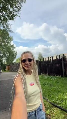The video is a selfie-style recording taken outdoors. A woman with blonde braids and sunglasses is walking on a wooden path. She is wearing a light yellow tank top and blue jeans. In some frames, a small child in a light-colored outfit can be seen walking ahead of her on the path. The environment appears to be a park or nature area with trees, greenery, and some wooden fences and signs. The sky is partly cloudy.