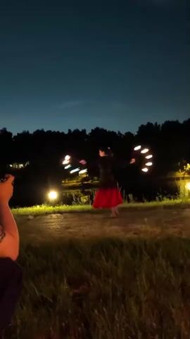 A person is spinning a prop with lights attached to it. The person is wearing a dark top and a red skirt. The background appears to be a park at night, with trees and lights visible. The sky is dark blue, indicating twilight or night. There is grass in the foreground. In the lower left corner, a person's arm holding a camera is visible, suggesting the video is being recorded. The lights on the prop create a circular effect as they are spun.