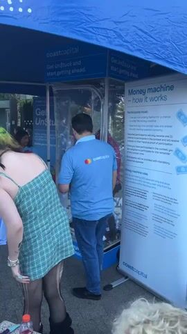The video opens on a close-up shot of the back of a blue t-shirt with the logo "coastcapital" printed in white. The logo features two overlapping circles, one red and one yellow, to the left of the text. The camera then pans slightly to the left and zooms out to reveal more of the scene. A person wearing the blue t-shirt, identified as a man, is standing in front of a clear plastic enclosure, possibly a money machine. He is facing away from the camera, looking into the enclosure. Inside the enclosure, there are what appear to be paper bills suspended in the air, possibly being blown by a fan, consistent with a money machine game. To the left of the man, a person with green dyed hair is partially visible, wearing a green and white checkered dress and black stockings. They are leaning forward and looking into the money machine. To the right of the man, a large white sign with text in black and blue is visible. The sign has the heading "Money machine - how it works" and lists rules and instructions for participants. In the background, other people and structures are dimly visible, suggesting an outdoor event. The lighting is bright, indicating daytime. The camera movement is subtle, with slight panning and zooming throughout the short clip.
