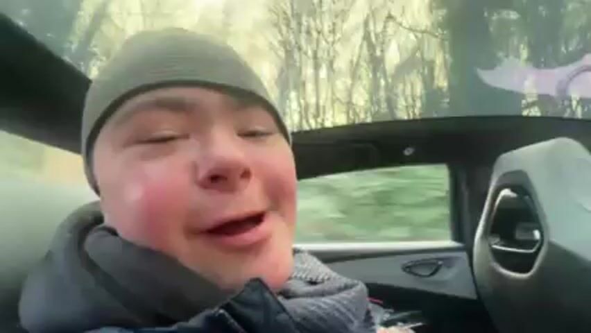 A young man wearing a beanie and jacket is seated in the backseat of a car. The camera is positioned directly in front of him. He smiles, sticks his tongue out slightly, and says "Merry Christmas." He then makes a kissing sound into the camera and smiles again. The car is moving, as the background shows trees passing by through the sunroof.