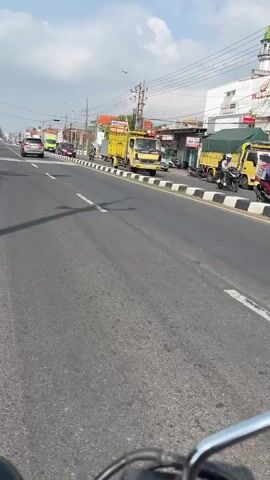 The video shows a daytime view from a vehicle moving along a paved road. The road has multiple lanes and is lined with businesses and buildings. Several vehicles are visible, including cars, trucks, and motorcycles. Some of the trucks are yellow and appear to be carrying cargo covered with tarps. The sky is partly cloudy, and there are utility poles with wires running alongside the road. Shadows of overhead structures, likely utility poles or streetlights, are cast across the road. The scene appears to be in a busy urban or suburban area.