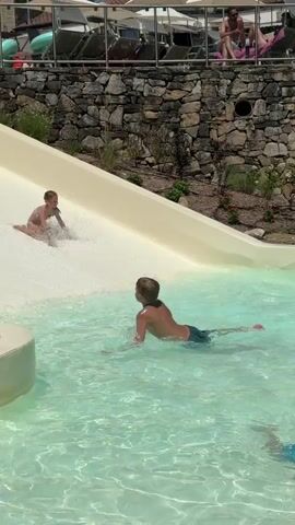 At 00:00, a child emerges from a water slide, creating a splash in a pool. Another child lies in the water nearby, facing away from the camera.  At 00:01, a young girl with dark hair in a swimsuit slides down the water slide. At 00:02, she is closer to the bottom of the slide. At 00:03, another child slides down, and the girl who was previously seen is now interacting with the water at the base of the slide. At 00:04, another child on the slide is partially submerged in water, while the child from the beginning of the clip is seen from behind. The pool is filled with clear, turquoise water. In the background, there is a stone wall, landscaping, and lounge chairs with people sitting on them.