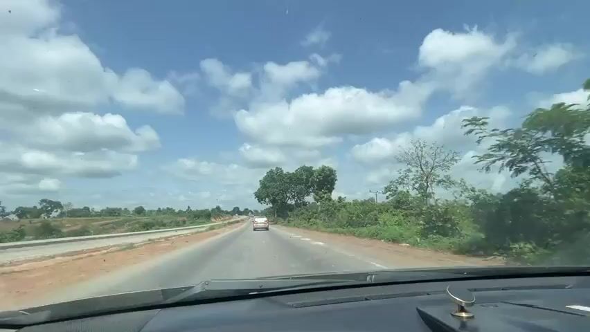 This video shows a view from inside a vehicle traveling on a paved road. The road has two lanes and a white line marking the center. There is a car ahead in the distance. The road is bordered by a dirt shoulder and sparse vegetation, including trees and bushes. To the left of the road, there is construction or unfinished roadwork. The sky is blue with scattered white clouds. The interior of the car is visible in the foreground, including the dashboard and a decorative item. The video appears to have been recorded during the daytime.