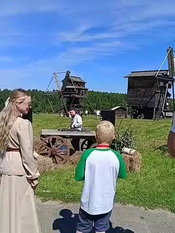 The video shows an outdoor event with several people and structures in the background. In the foreground, facing away from the camera, is a young boy wearing a green and white shirt. To his left, also facing away from the camera but turned slightly towards the viewer, is a woman with long blonde hair wearing a light-colored dress and a large white bow in her hair. Between them, a man in a white shirt is standing behind a wooden cart with large wheels, appearing to operate some equipment on it. In the background, there are several wooden structures that resemble old buildings or windmills, and a dense line of trees. The sky is clear blue with scattered clouds. The ground is covered in green grass and hay bales. The overall impression is of a historical reenactment or cultural festival.