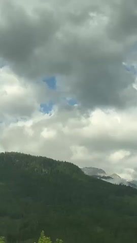 This media file captures a scenic view of a mountainous landscape, likely in Bischofshofen, Austria. The scene is dominated by lush green forested slopes in the foreground, rising towards more rugged, snow-capped peaks in the distance.

The sky is filled with dramatic, heavy clouds, ranging from dark grey to bright white, with patches of blue sky peeking through. This suggests a dynamic weather pattern, possibly transitioning between rain and sun. The lighting is diffused, indicating it might be a cloudy day, possibly late afternoon or early morning, though it's difficult to determine the exact time of day.

In the mid-ground, there are hints of human habitation with small buildings and cultivated fields visible nestled amongst the hills. The overall emotion evoked is one of natural grandeur and perhaps a sense of peacefulness, despite the somewhat ominous clouds. There is no visible text within the media file. The activity is simply the observation of this natural landscape, suggesting a view from a moving vehicle or a stationary viewpoint.