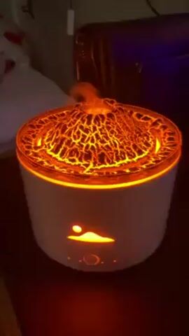 The video shows a white, cylindrical object emitting orange light. The top of the object is textured and resembles lava, with cracks and fissures illuminated by the light. There is also an orange light shining through a design on the front of the object, which appears to be a depiction of a mountain with a sun or moon above it. There are also a few buttons or touch sensors on the front of the object. In the background, there is a bed with patterned bedding, a dark-colored suitcase or bag, and other objects that are less visible. The video appears to be taken in a dimly lit room.