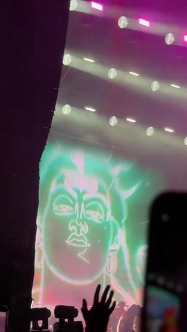 A low-angle video shot from within a crowd at a concert shows a large screen displaying an image of a person with a snake around their neck, overlaid with a green misty effect. Above the screen, pink lights illuminate the stage and a sign that reads "PARK LIVE." The crowd is visible in the foreground, with many people holding up their phones to record the event. Some audience members are raising their hands in the air. The lighting is predominantly pink and purple, with some white spotlights. The overall atmosphere is energetic and engaging.