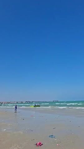 The video opens to a wide shot of a beach with a clear blue sky and turquoise ocean waves lapping at the shore. The beach is sandy and appears to have some debris scattered on it. As the video progresses, the camera pans to the right, revealing two fishing boats docked on the sand. The boats are green and white, with fishing equipment visible. The scene is peaceful, with no people or animals present.