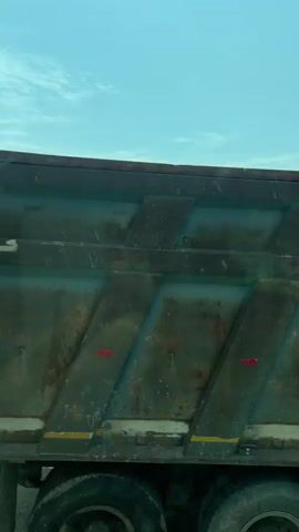 The video shows a view from a moving train.  In the initial frame, a man wearing a turban and light-colored shirt operates a small blue machine next to what appears to be a pile of railroad ties. The area looks like a roadside or rail yard with a shed-like structure in the background.  The video then transitions to a view of trees and vegetation passing by the train window against a blue sky. The camera appears to be focused on the nearby foliage as it moves along.