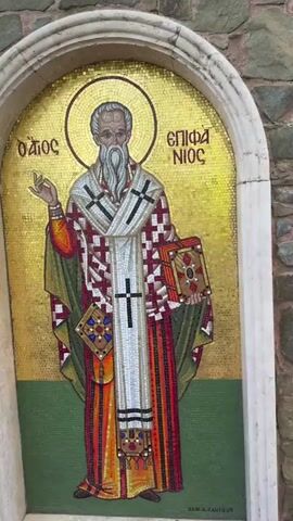 The video begins with a close-up of a mosaic of Saint Nicholas on a stone wall. The camera then pans to the right, showing more of the stone wall which appears to be part of an exterior building. The wall is constructed of irregularly shaped stones with mortar in between. There are arched recesses in the wall. The camera continues to pan right, revealing a red brick pathway with a black and white striped border. On the left side of the pathway is a low stone wall topped with a white coping. Beyond the stone wall is lush green foliage. On the right side of the pathway, the stone wall continues, also with arched recesses. Two people in white clothing are walking away from the camera down the pathway. The scene appears to be outdoors in daylight.