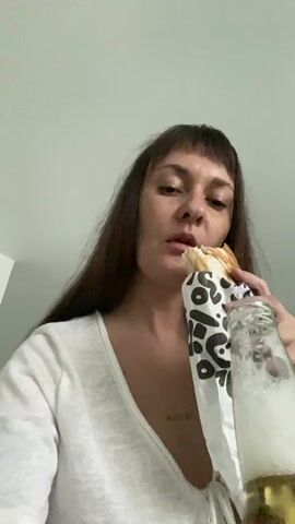 This media file shows a close-up shot of a woman eating and drinking. She has long, dark brown hair with bangs and is wearing a white v-neck top. She is holding a burrito wrapped in white paper with a black leopard-like print pattern in her right hand and a clear glass bottle of beer in her left hand. The beer appears to be a light lager, with a foamy head. The woman is looking directly at the camera with a neutral expression, and appears to be taking a bite of the burrito while holding the beer. The background is a plain, light-colored wall. There is a small tattoo on her chest, which appears to read "mirror". The time of day is not discernible, nor is the weather. The setting appears to be indoors, possibly a home.