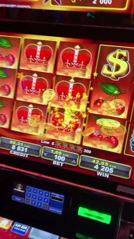 The video shows a close-up of a slot machine with bright, flashing lights and animated symbols. The reels are in motion, displaying symbols such as cherries, crowns, gold coins, and a dollar sign. There is a digital display showing credit, bet, and win amounts in Euros. Below the main screen, there is a keypad and a touch screen with betting options and game selection buttons. The overall environment appears to be a casino or gaming venue, suggested by the ambient lighting and the nature of the machine. The perspective suggests someone is operating the machine and recording the gameplay.