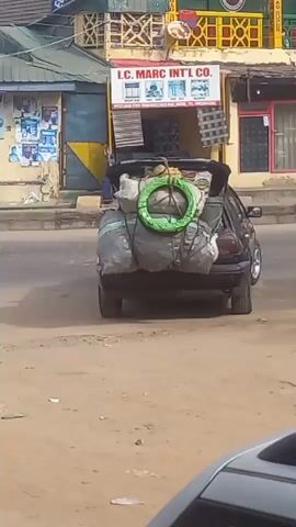 The video shows a black car with a heavily loaded roof rack and trunk, parked on a street in what appears to be a West African town. The car is packed with large bundles wrapped in gray tarps and tied with rope, with a green ring-shaped object on top. Nearby, two women and two men are standing. One of the women wears a black hijab and a white and green patterned dress. The second woman wears a beige top and a black skirt. One of the men is seated in the back of a parked silver car. The other man is wearing a hat. Buildings with signs are visible in the background. A white sedan is parked behind the black car.  The ground is dusty.  The footage seems to be taken from inside another car.