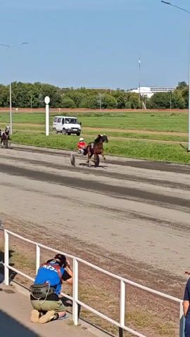 The video shows a horse race on a dirt track. In the foreground, two people are seated, watching the race. A third person, a photographer, is kneeling and taking pictures. The track is surrounded by a grassy area and trees. In the background, there are several flags and a white van. The horses are pulling sulkies, which are lightweight racing carts. The horses are running at full speed, and the drivers are leaning forward in their seats. The video captures the excitement and intensity of the race.